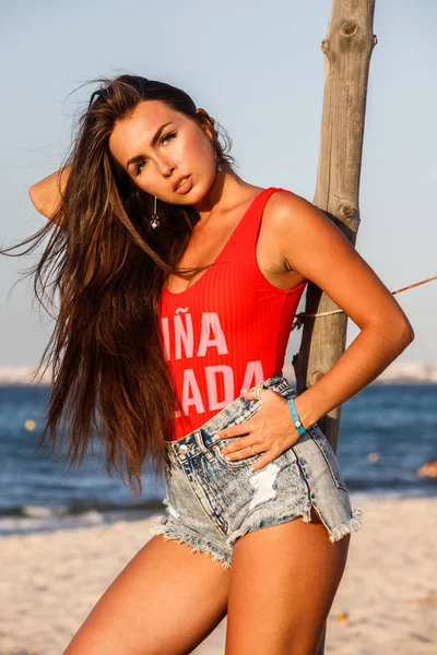 美丽的黑发模型在红色泳衣和蓝色牛仔裤短裤站在沙滩上白天 — 图库照片
