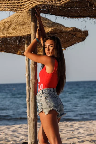 美丽的黑发模型在红色泳衣和蓝色牛仔裤短裤站在沙滩上白天 — 图库照片