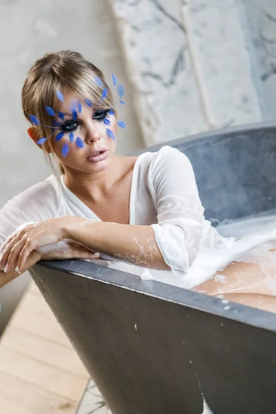 浴室を取っている白い服の美しい金髪の女性 顔に小さな青い羽を持つスタイリッシュな化粧品で美容モデルの肖像 — ストック写真