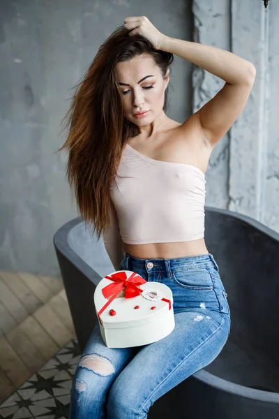 Portrait of beautiful young woman in blue jeans and beige t-shirt. Young model sitting on bath with box of sweets in hands, luxury apartments interior background