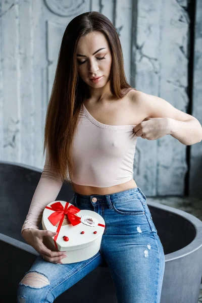 Portrait of beautiful young woman in blue jeans and beige t-shirt. Young model sitting on bath with box of sweets in hands, luxury apartments interior background