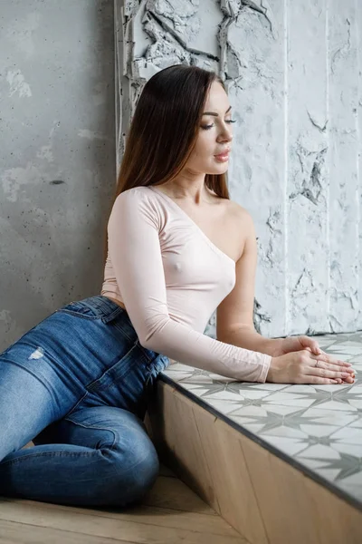 穿着蓝色牛仔裤和米色 T恤的美丽年轻女子的肖像 坐在豪华公寓地板上的年轻模特 — 图库照片