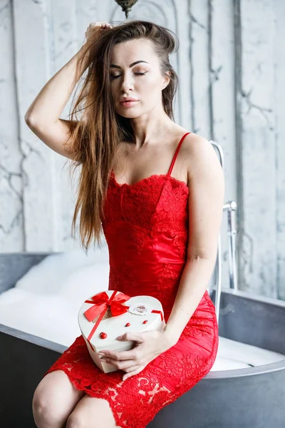 赤いドレスの美しい若い女性の肖像画 箱のお菓子付きのバスルームに座っているモデルします — ストック写真