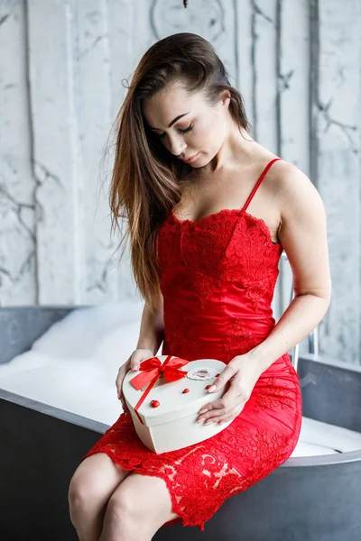 Portrait of beautiful young woman in red dress. Model sitting in bathroom with box of sweets
