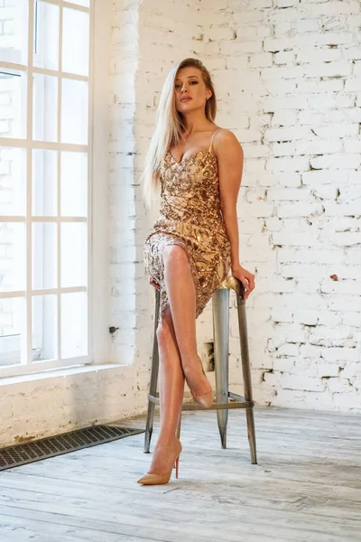 スタジオ 白いレンガの壁の背景に立ってスタイリッシュな黄金のドレスの美しさのブロンドの女性のファッションポートレート — ストック写真
