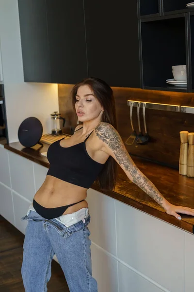 毛茸茸的黑发女人 手上有纹身 在厨房里摆姿势 穿着蓝色牛仔裤和黑色上衣站在公寓内部的年轻女子的画像 — 图库照片