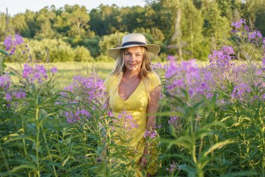 Yazlık giysiler içinde sarışın güzel bir kadının açık hava portresi. Gündüz vakti çiçek tarlasında poz veren şapkalı ve sarı elbiseli genç bir model. 