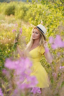Yazlık giysiler içinde sarışın güzel bir kadının açık hava portresi. Gündüz vakti çiçek tarlasında poz veren şapkalı ve sarı elbiseli genç bir model. 