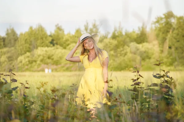 夏服の美しさブロンドの女性の屋外肖像画 昼間の花畑でポーズをとる帽子と黄色のドレスの若いモデル — ストック写真