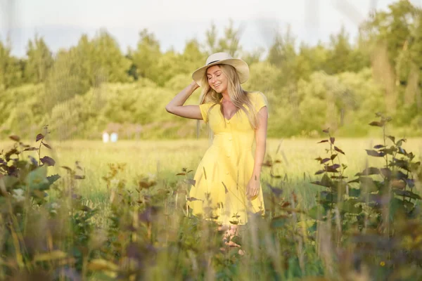 夏服の美しさブロンドの女性の屋外肖像画 昼間の花畑でポーズをとる帽子と黄色のドレスの若いモデル — ストック写真