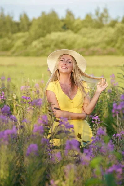 夏服の美しさブロンドの女性の屋外肖像画 昼間の花畑でポーズをとる帽子と黄色のドレスの若いモデル — ストック写真