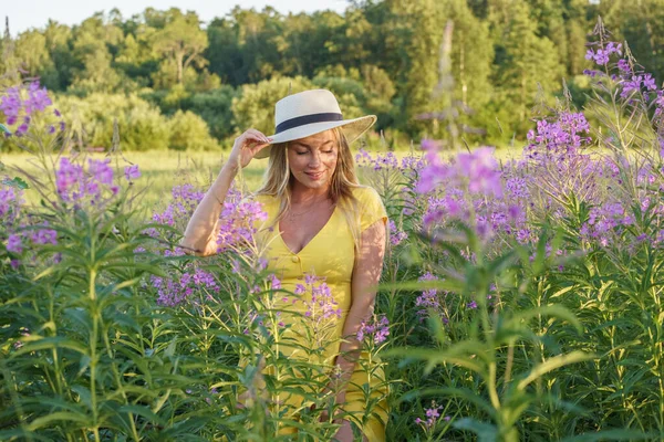 夏服の美しさブロンドの女性の屋外肖像画 昼間の花畑でポーズをとる帽子と黄色のドレスの若いモデル — ストック写真