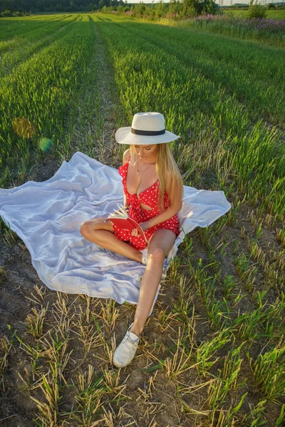 夏服の美しさブロンドの女性の屋外肖像画 昼間は花畑で休む帽子と赤いドレスの若いモデル — ストック写真