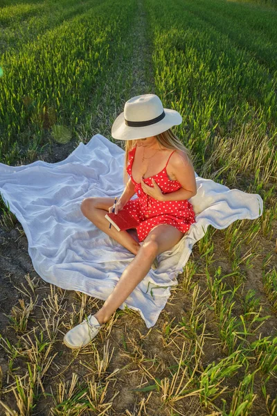 夏服の美しさブロンドの女性の屋外肖像画 昼間は花畑で休む帽子と赤いドレスの若いモデル — ストック写真