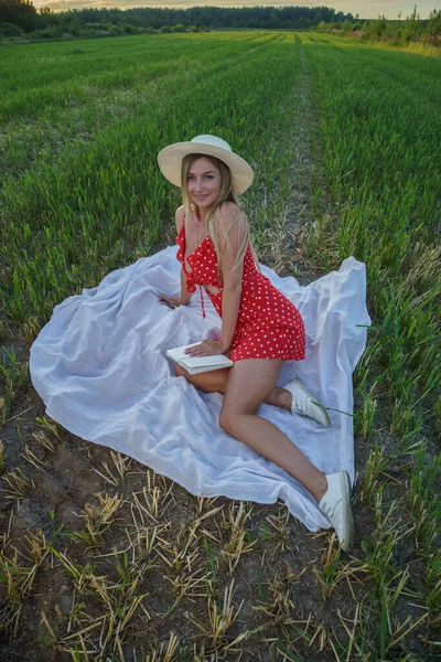 夏服の美しさブロンドの女性の屋外肖像画 昼間は花畑で休む帽子と赤いドレスの若いモデル — ストック写真