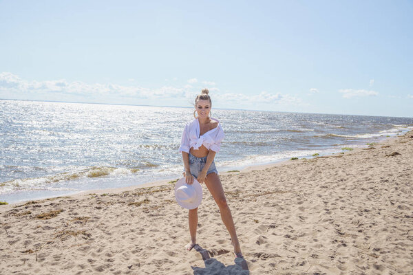 На улице портрет красотки блондинки в белой рубашке и синих джинсовых шортах. Позирование модели на пляже