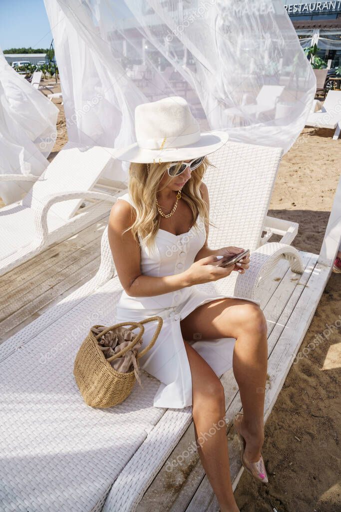 Summer portrait of beauty blond woman in white dress and white hat. Model resting in lounger on beach