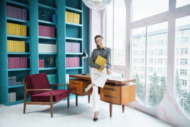 Güzel genç bir kadın kütüphanenin içinde poz veriyor. Gayri resmi giyinmiş esmer bir kadın portresi. 