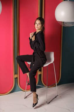 Lüks dairelerde kırmızı duvara yakın poz veren güzel bir iş kadını. Siyah takım elbiseli ve yüksek topuklu güzel bir esmer portresi. 