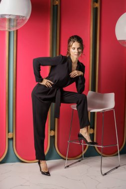 Lüks dairelerde kırmızı duvara yakın poz veren güzel bir iş kadını. Siyah takım elbiseli ve yüksek topuklu güzel bir esmer portresi. 