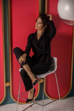 Lüks dairelerde kırmızı duvara yakın poz veren güzel bir iş kadını. Siyah takım elbiseli ve yüksek topuklu güzel bir esmer portresi. 
