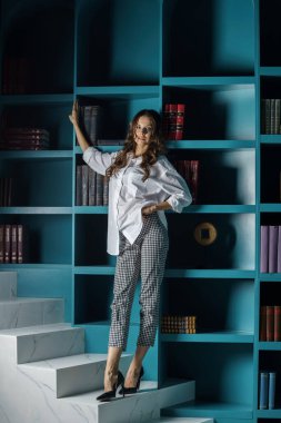 Güzel genç bir kadın kütüphanenin içinde poz veriyor. Gayri resmi giyinmiş esmer bir kadın portresi. 