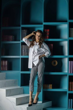 Güzel genç bir kadın kütüphanenin içinde poz veriyor. Gayri resmi giyinmiş esmer bir kadın portresi. 