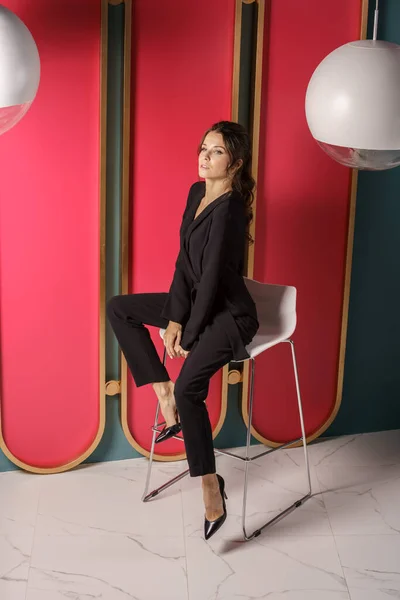 Beautiful businesswoman posing close to red wall at luxury apartments interior. Portrait of beauty brunette in black suit and high heels