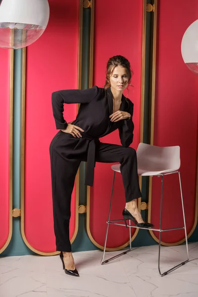 Beautiful businesswoman posing close to red wall at luxury apartments interior. Portrait of beauty brunette in black suit and high heels