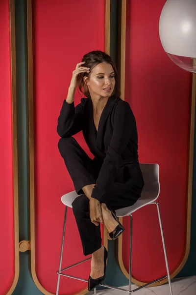 Beautiful businesswoman posing close to red wall at luxury apartments interior. Portrait of beauty brunette in black suit and high heels