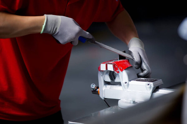 Человек в красной футболке и серых перчатках обрабатывает металлическую деталь, зажатую в тиски, которая устанавливается на рабочем столе в гараже
