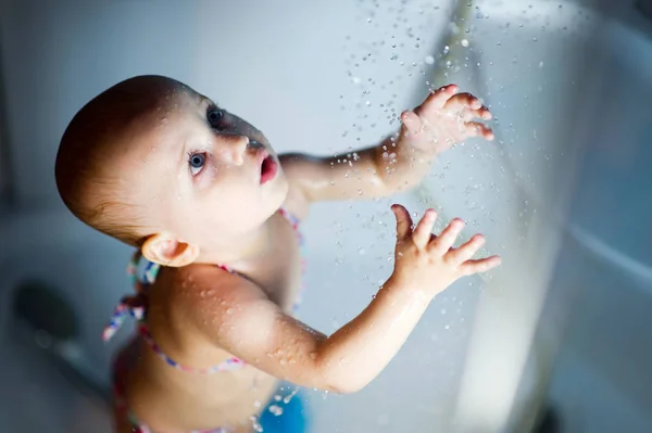 Ein kleines Mädchen im Badeanzug steht unter der Dusche, blickt auf das strömende Wasser und greift danach — Stockfoto