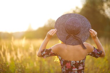 Büyük kenarlı koyu şapkalı bir kız kameraya sırtını döner ve gün batımına bakar..