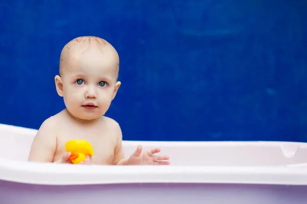 Маленька дитина грає з жовтою качкою в білій ванні на синьому фоні — стокове фото