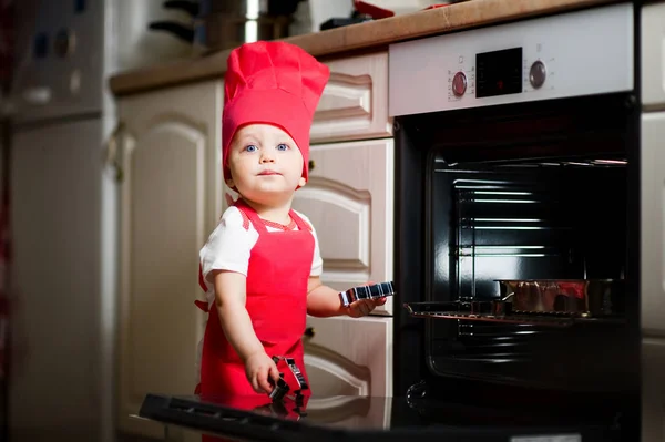Dziewczynka w czerwonym stroju kucharza odgrywa z naczynia kuchenne w pobliżu pieca. Bawią się w kuchni z bliska — Zdjęcie stockowe