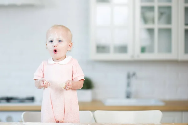 Das kleine Mädchen mit Mehl im Gesicht und Teigscheiben in den Händen steht hinter der Arbeitsplatte in der Küche und blickt in den Rahmen, öffnet den Mund, schneidet. — Stockfoto