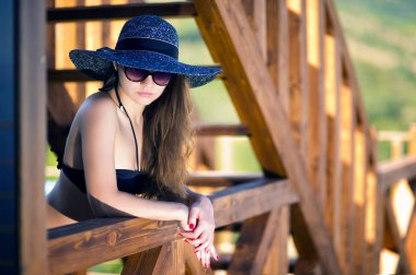 siyah mayo, güneş gözlüğü ve şapkalı bir kız ahşap bir çitin yanında duruyor.