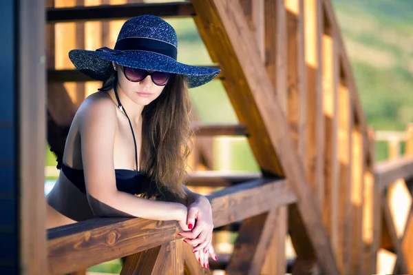 siyah mayo, güneş gözlüğü ve şapkalı bir kız ahşap bir çitin yanında duruyor.