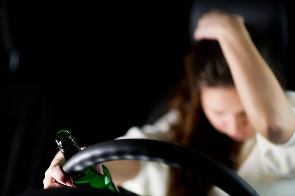 Condução bêbada. uma mão com uma garrafa de álcool está no volante, e no desfoque a menina bêbada segura a cabeça — Fotografia de Stock