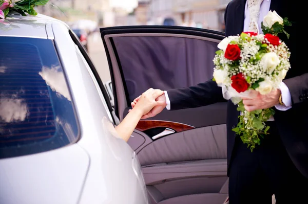 Le marié avec un bouquet à la main donne sa main à la mariée quand elle sort de la voiture — Photo