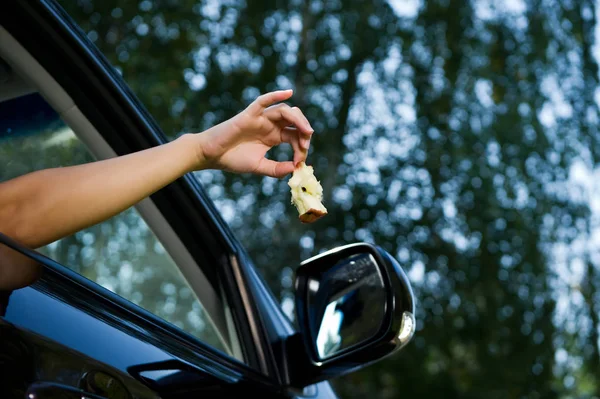 Jeune femme tient dehors et est sur le point de jeter un noyau de pomme par la fenêtre de la voiture ouverte. Vue du bas, sur fond d'arbres flous et de ciel. Gros plan — Photo