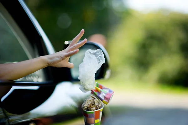 Egy fiatal nő dob hulladékot az autó-egy kávéscsésze és egy táska. Környezetszennyezés, szelektív fókusz, háttérvilágítás — Stock Fotó
