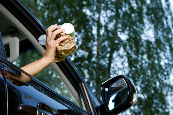 The girl is going to throw a handful of garbage accumulated in the car into the open window of the car. Bottom view, against the background of blurry trees and sky, summer day