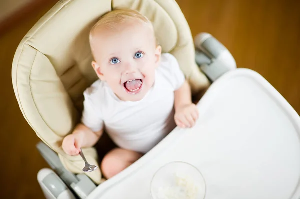 Ребенок в детском кресле для кормления ест кашу и смотрит вверх, радостно улыбаясь — стоковое фото