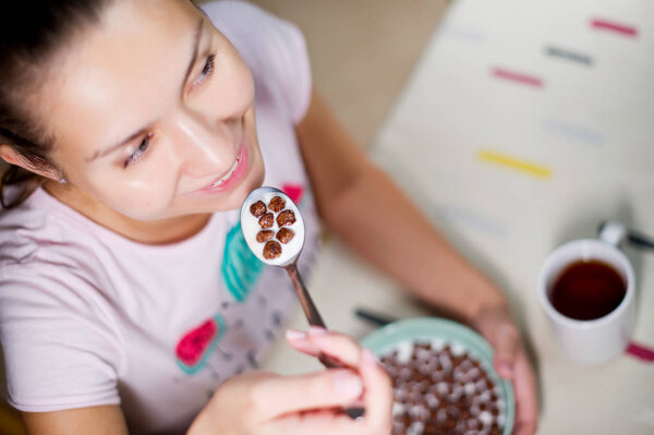 smiling girl holding a spoon with chocolate balls and milk near the mouth over the table