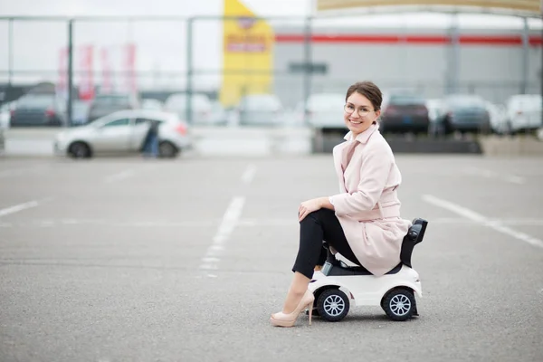 Junge Frau mit Brille auf einem Supermarkt-Parkplatz, die auf einem Plastikauto für Kinder sitzt und lächelt — Stockfoto