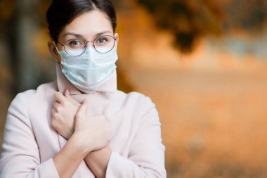 Tıbbi maskeli ve gözlüklü bir kız hastalık ve soğuk sırasında ceketinin yakasını daha sıkı kapatır. Yüz maskeli kadın. Kapat, boşluğu kopyala