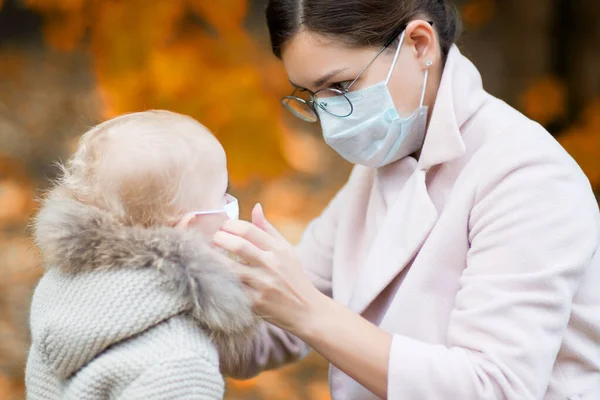 Tıbbi maskeli bir anne, sonbaharda bahçede olan küçük kızına tıbbi maskeyi ayarlar. — Stok fotoğraf