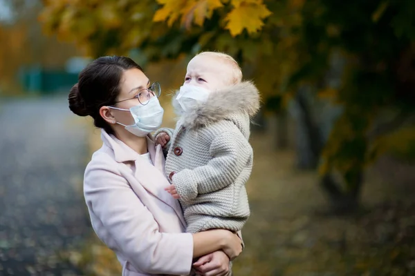 Tıbbi maskeli bir anne, ağlayan bir kızı kollarında tutuyor ve ona bakıyor. — Stok fotoğraf