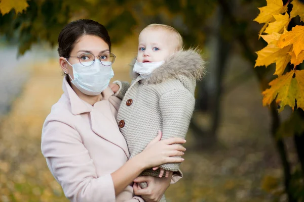 Mutter mit medizinischer Gesichtsmaske hält eine kleine Tochter im Arm, vor dem Hintergrund eines herbstlichen Waldes — Stockfoto